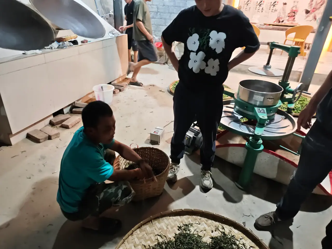 People processing fresh tea at night beside woks, bamboo trays and a rolling machine